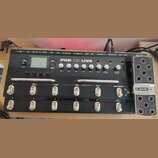 Černý kytarový procesor Line 6 POD X3 LIVE s kovovým pedálem vpravo. Na horním panelu jsou ovládací knofíky s nápisy DRIVE, BASS, MID, TREB, PRES, VE…