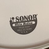 Detail bílého bubnového koženě s černým potiskem. Je na něm vidět logo SONOR a nápisy White Medium, DRUM HEAD, MADE BY REMO a U. S. A. Povrch je hlad…