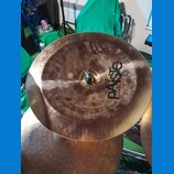 Kruhová činely v bronzové barvě s viditelnými stopy opotřebení a nečistotami na povrchu. Na činely je černý nápis pAiSte a pod ním menší text MADE IN…