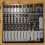 Stříbro-šedý mixážní pult značky behringer s nápisem XENYX X1222USB. Zařízení obsahuje 6 mikrofonních vstupů, několik linkových vstupů a faderové ovl…