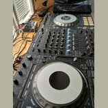 Černý DJ 컨트롤러 s ovládacími prvky, posuvníky a dvěma stříbrnými jog-wheels. Na povrchu je viditelný nápis Pioneer DJ. Zařízení vykazuje běžné známky p…
