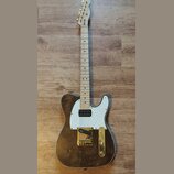 Elektrická kytara typu Telecaster s hnědým dřevěným povrchem těla a bílým perleťovým pickguardem. Nástroj má zlatý hardware, včetně ladíček a mostu,…