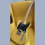Žlutá elektrická kytara s tvarem typu Telecaster a černým pickguardem. Na hlavě hředu je viditelné logo Fender. Tělo kytary vykazuje výrazné opotřebe…