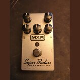 Stříbrný efekt pedál s pěti černými ovládacími knoflíky. Na přední straně je umístěno logo MXR, nápis Super Badass a pod ním slovo DISTORTION. V horn…