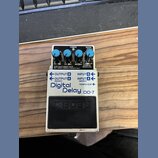 Stompbox efekt s bílým kovovým šasi a černým spínacím pedálem. Na přední straně je nápis Digital Delay DD-7 a logo BOSS. Zařízení má čtyři modré ovlá…
