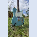 Světle modrá elektrická kytara typu Telecaster s bílým pickguardem a hnedým hmatníkem. Nástroj je umístěn na černém stojanu v trávě. Na kytare nejsou…