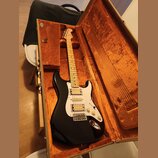 Černá elektrická kytara v hnědém tvrdém kufru s oranžovou výplní. Nástroj má bílý pickguard, tři snímače a světle hnědý javorový hриф. Na hlavě kytar…