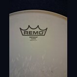 Detail bílého, matného bicího koženu. Uprostřed je černé logo REMO v podobě koruny a pod ním nápisy EMPEROR®, COATED a MADE IN U. S. A. V dolní části…