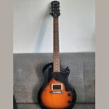 Elektrická kytara s tělem v žlutém sunburst provedení a černým pickguardem. Na hlavě hředu je bílý nápis Epiphone a pod ním nápisy Junior a Les Paul.…