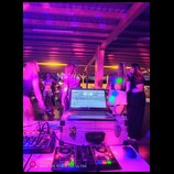 Fotografie zachycuje DJské vybavení v provozu na akci. Na bílém transportním kufru stojí otevřený notebook značky hp, pod ním je umístěn DJ kontrolér…