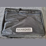 Černý textilní pouzdro na hudební nástroj s dvěma rukojtěmi a zipem. V dolní části je bílý obdélníkový štítek s nápisem ROCKBOARD® a pod ním menší te…
