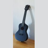 Černý ukulele s matným povrchem. Na hlavě nástroje je bílý nápis „The Waterman USA“. Nástroj vykazuje známky běžného používání a je opřen o bílou zeď…
