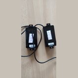 Dvě černé elektrické snímače s připojenými kabely. Na levém je nalepený bílý papírek s červeným nápisem „KRY“ a štítek s textem „L 3N-BLK M 002MTM N“…
