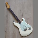 Bílá elektrická kytara s jasně viditelným logem Fender na hlavě. Nástroj má tmavý hmatník, světle zelený pickguard a tři snímače. Povrch kytary vypad…