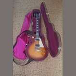 Elektrická kytara s tónem sunburst v hnědých a žlutých odstínech, bílým pickguardem a dvěma humbuckery. Nástroj je umístěn v otevřeném hnědém tvrdém…