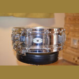 Kovový snare drum v chromovaném provedení. Na korpusu je vidět oválný štítek s nápisy DYNA-SONIC, SERIAL No. (číslo nečitelné) a CUSTOM BUILT. Na mec…