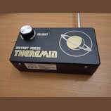 Černý obdélníkový přístroj s nápisem DISTANT VOICES THEREMIN a grafikou planety s prstencem. Na horní straně je stříbrný ovládací knoflík a nápis ADJ…