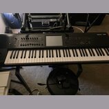 Černý digitální klavír umístěný na černém stojanu. V pravém horním rohu je bílý nápis KRONOS Xsynth. Nástroj má v horní části ovládací prvky, tlačítk…