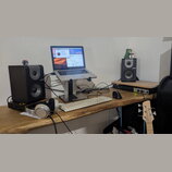 Na dřevěném stole je umístěno studio s notebookem na stojanu, dvěma černými studiovými monitory s nápisem M-Audio a bílou klávesnicí. V popředí leží…