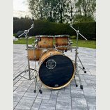 Akustická bicí souprava z přírodního dřeva s kovovým hardwarem. Na basovém bubnu je černé logo s nápisem „SINCE 2003 DC DRUMS handcrafted in germany“…