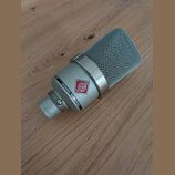 Stříbrný kondenzátorový mikrofon s kovovou síťovkou. Na těle je nápis NEUMANN MICROPHONE a červenobílý štítek s nečitelným nápisem. Na spodní části j…