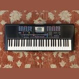 Černý digitální klavír s bílými a černými klávesami. Na levé straně panelu je nápis YAMAHA a vpravo označení modelu PSR-220. Horní část přístroje obs…
