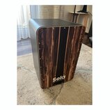 Černý cajon s přední deskou v imitaci tmavého dřeva. Ve spodní části je bílý nápis Sela a pod ním menší text the soul of sound. Povrch je hladký, v r…