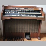 Hnědý dřevěný orgán se dvěma klávesnicemi a pedálovým hracím polem. Na spodní klaviatuře je umístěn nápis HAMMOND. Horní panel obsahuje řadu bílých a…