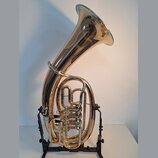 Zlatavý mosazný tuba v černé stojce. Na vnitřní straně zvonu je vidět zlatý nápis "Yamaha". Nástroj vykazuje známky běžného používání v podobě drobný…