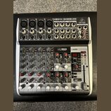 Šedý mixážní pult značky behringer s nápisem XENYX QX1202USB. Zařízení obsahuje několik vstupních kanálů s ovládacími prvky pro GAIN, EQ a FX, USB IN…