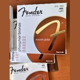 Fotografie zobrazuje tři krabičky strun pro akustickou kytaru. Na obalech je bílé pozadí, hnědý kruh se zlatým logem „F“ a nápisy „Fender“, „MAKE HIS…