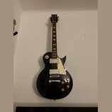 Černá elektrická kytara s tvarem Les Paul, bílým pickguardem a dvěma humbuckery. Na hlavě hnedku je viditelné logo Epiphone. Nástroj vykazuje běžné z…