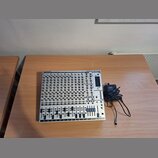 Stříbrný mixážní pult s černými posuvníky a ovládacími prvky. V horní části je uveden nápis BEHRINGER a model EURORACK MX 2642A. Vedle přístroje leží…