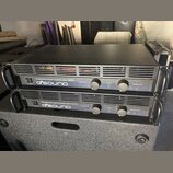 Dvě černé rackové konce s nápisem Professional Power Amplifier QSA-600 a logem QSOUND. Každý přístroj má v přední části vypínač, dva ovratové ovládac…