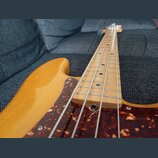 Elektrická basová kytara s přírodním dřevěným povrchem těla a hmatníkem. Nástroj má tmavě červený plexiglasový pickguard s mramorovaným vzorem. Na fo…
