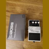 Na fotografii je kytarový pedál Boss Noise Suppressor NS-2 v bílém provedení s černým spínacím tlačítkem. Vedle pedálu je umístěna jeho originální še…