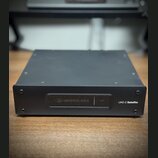 Černý kovový přístroj obdélníkového tvaru. Na přední straně je nápis UAD-2 Satellite a panel s logem a názvem UNIVERSAL AUDIO oraz nápisem HOST. Povr…