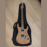 Elektrická kytara s přírodním dřevěným povrchem těla a hnedým hmatníkem, položená na černém softcasu. Nástroj má dva humbuckery a na hlavě je viditel…
