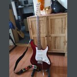 červená elektrická kytara s bílým pickguardem a hnedým hmatníkem. Na hlavě je viditelný nápis YAMAHA. Kytara je umístěna na stojanu a má připojený če…