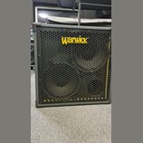 Černý basový reproduktor v šedém rámu s kovovou ochrannou mřížkou. V horní části je žluté logo Warwick. Za mřížkou jsou vidět dva velké reproduktory…