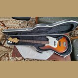 V otevřeném černém kufru s vnitřním čalouněním leží elektrická basová kytara. Nástroj má tělo v barvě sunburst, bílý pickguard a hnědý hmatník. Na hl…