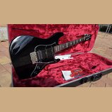 Černá elektrická kytara s tvarem typu Stratocaster v otevřeném kufru s červeným vnitřkem. Na hlavě kytary je bílý nápis Ibanez. Kytara má chromovaný…