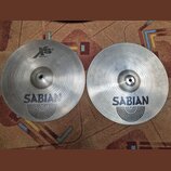 Dvě stříbrné kovové činely značky SABIAN s koncentrickým žilováním povrchu. Levý činele má v horní části logo XS, nápis 20 a text Hata W736 cm. Oba n…