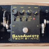 Černý efekt pedál s nápisy SansAmp GT2 a TECH 21 NYC. Na horní straně jsou čtyři ovládací knoflíky s popisky LEVEL, HIGH, LOW a DRIVE. Pod nimi se na…