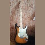 Elektrická kytara s tělem v žlutě-černé barvě (sunburst) a bílým pickguardem. Na hlavě hnedku je viditelné logo Squier. Hmatník je vyroben ze světléh…