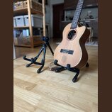 Dřevěný akustický ukulele s výřezem v těle, umístěné na černém stojanu. Vedle něj stojí druhý volný černý stojan. Na těle nástroje není vidět žádné l…