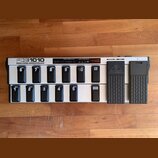 Stříbrný MIDI kontrolér s černými spínači a dvěma expresními pedály. Na horní straně je nápis FCB1010 MIDI FOOT CONTROLLER a na jednom z pedálů logo…
