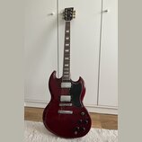 Kytara s tmavě červeným lakem a černým pickguardem. Na hlavě je viditelné zlaté logo Epiphone. Nástroj má dva humbuckery, tři ovládací knoflíky a pře…