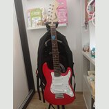 Červená elektrická kytara s bílým pickguardem a hnědým hmatníkem. Na hlavě nástroje je viditelné logo Squier. Kytara stojí na stojanu před černým bat…