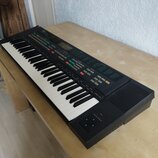 Černý elektronický klavír s bílými a černými klávesami. Na horním panelu jsou umístěny barevné ovládací tlačítka, displej a nápisy, z nichž je čiteln…
